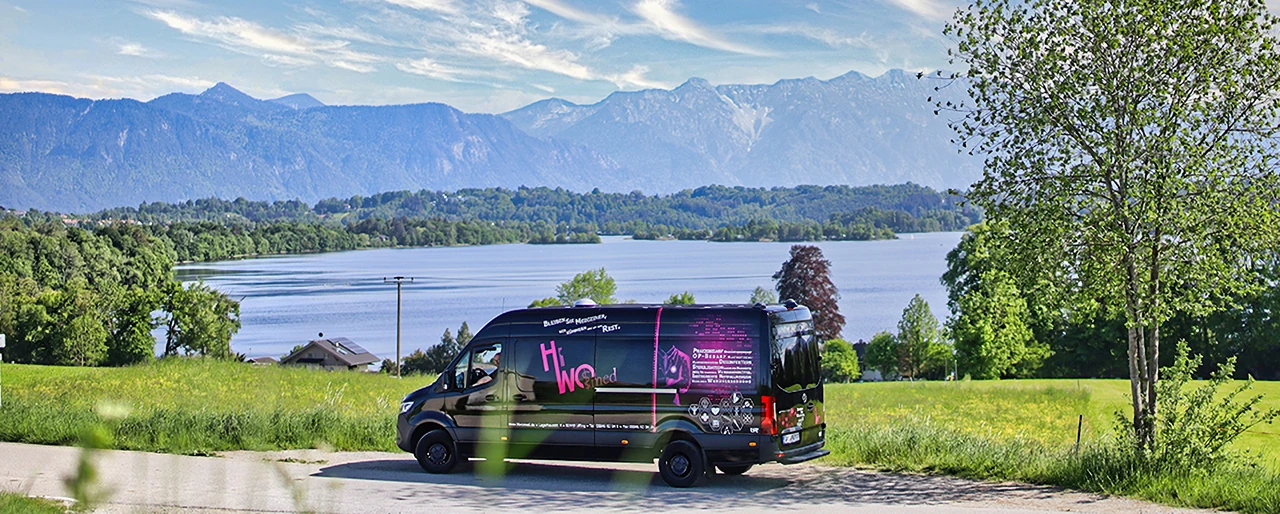 Ein HiWo-med-Transporter auf einer Straße am Staffelsee, dahinter die Alpenkette.