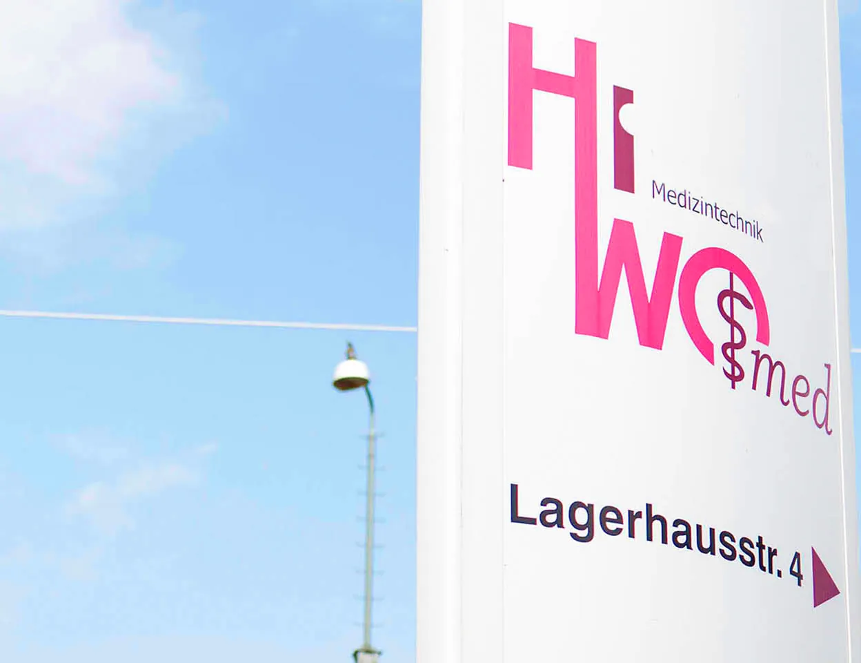 Das Firmenschild von HiWo-med mit Logo und dem Hinweis „Lagerhausstr. 4“ vor blauem Himmel.